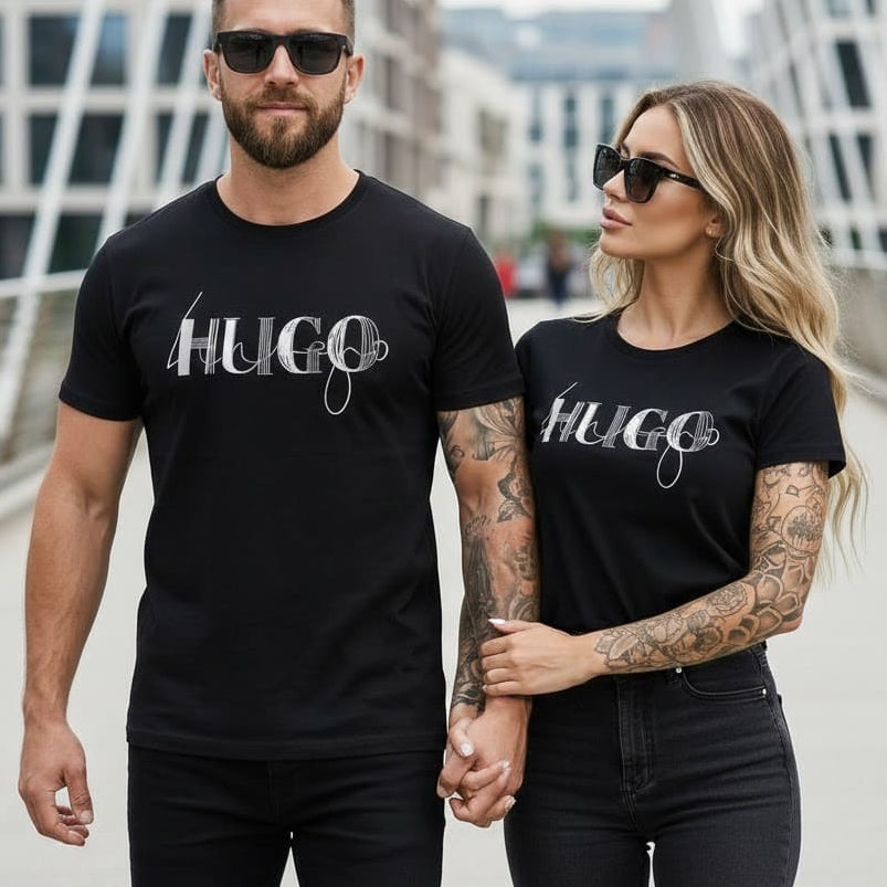Мъж и жена с еднакви черни тениски Hugo с сребърно графично лого на мост в града.