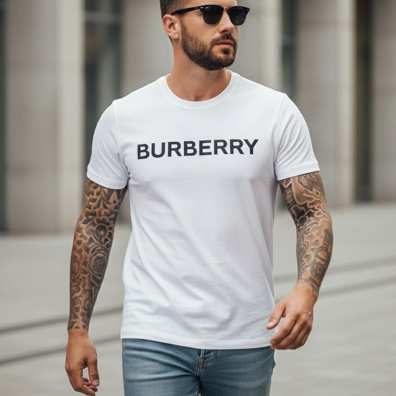 Мъж с татуировки по ръцете, облечен в бяла тениска с черен надпис Burberry и сини дънки.