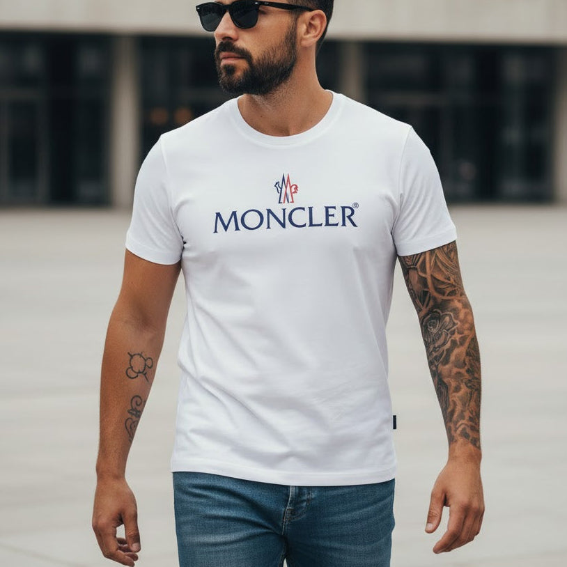 Мъж със слънчеви очила и татуировки, облечен в бяла тениска с лого на Moncler и дънки на открито.
