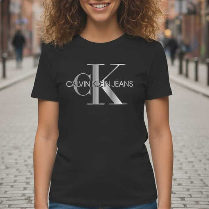 Усмихната жена с къдрава коса, носеща черна дамска тениска Calvin Klein Jeans с голямо сребристо лого на градски фон.